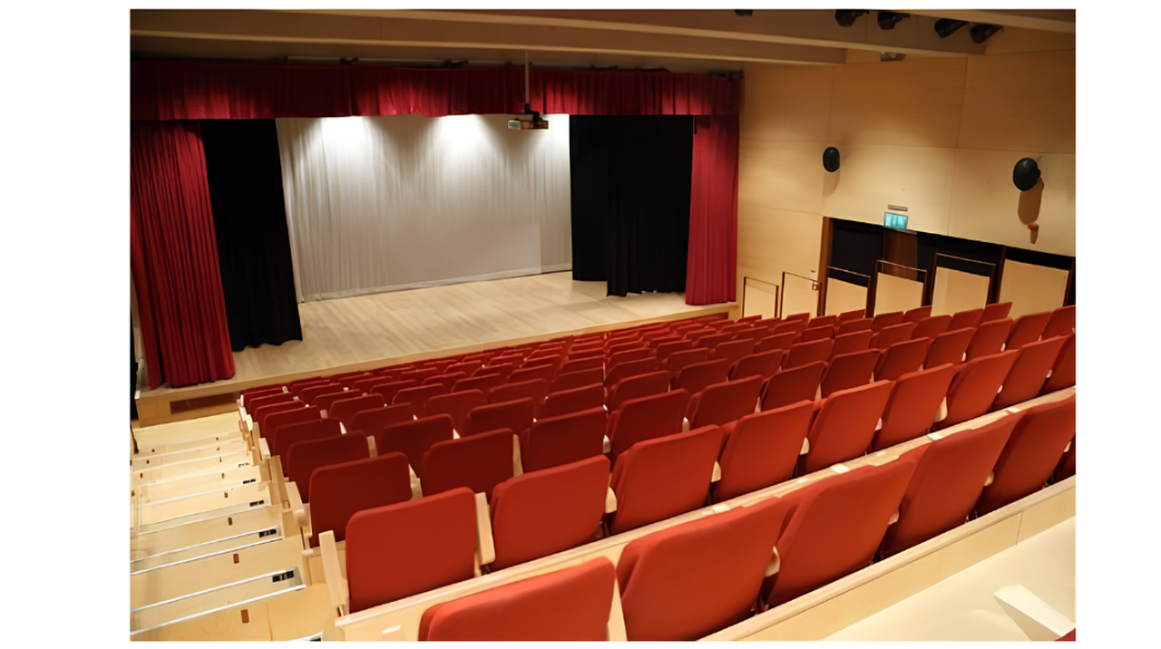 Empty auditorium with red seats and a wooden stage. The stage is framed by red curtains and has a light gray backdrop.